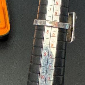 May include: A silver ring with a dark blue stone set in a square bezel. The ring is being sized on a metal ring sizer with markings from 6 to 10.