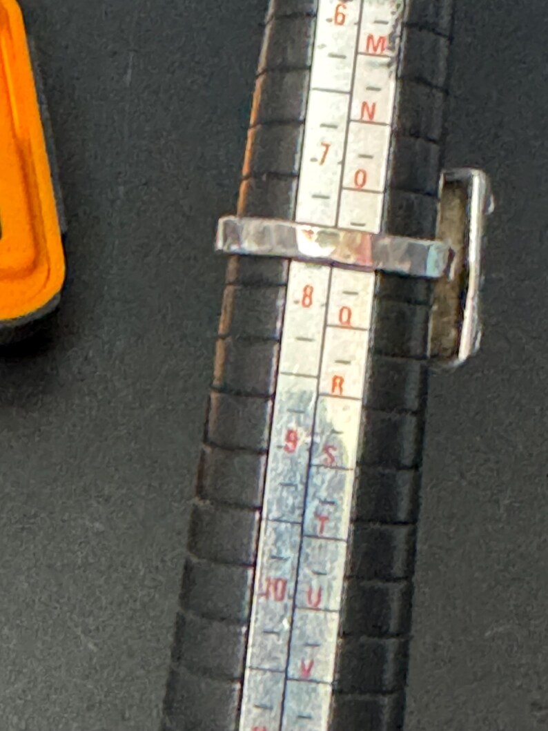 May include: A silver ring with a dark blue stone set in a square bezel. The ring is being sized on a metal ring sizer with markings from 6 to 10.