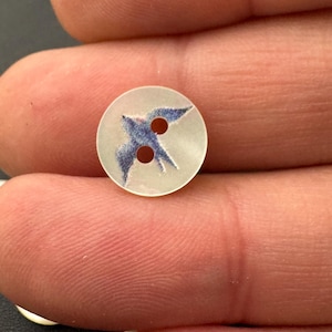 Pu&ograve; includere: Un piccolo bottone rotondo con finitura traslucida e madreperlacea. Il bottone presenta un'illustrazione blu e bianca di un uccello in volo. Due piccoli fori sono visibili per cucire. Questo bottone &egrave; ideale per progetti di cucito.