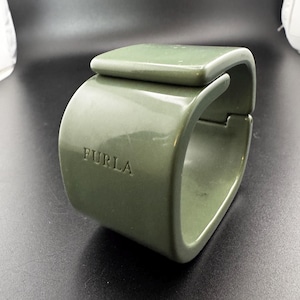May include: A sage green cuff bracelet with a smooth, glossy finish and a wide, structured band. The word FURLA is embossed on the side of the bracelet. The design features a rectangular top section that overlaps the main body of the cuff.