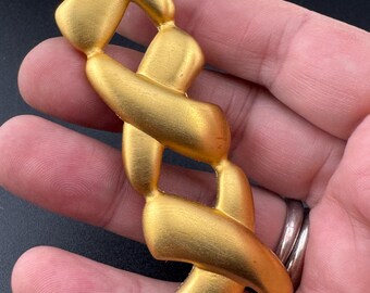 May include: A gold-toned metal brooch with a woven design. The brooch is shaped like a long, thin rectangle.
