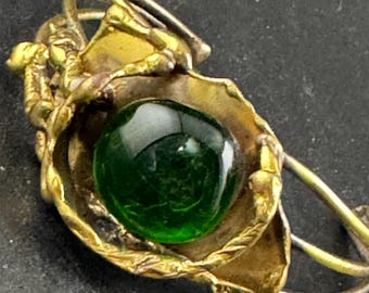 May include: A vintage brooch featuring a vibrant green glass cabochon set within a gold-toned metal floral design. The brooch has a detailed, organic aesthetic, with textured leaves and delicate metalwork. The pin is set against a dark background.