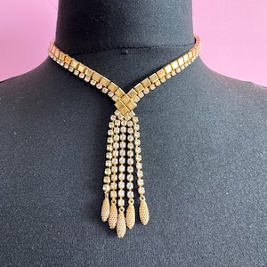 May include: A gold-toned necklace with a square and rectangular design, adorned with clear rhinestones. The necklace features a Y-shaped pendant with dangling strands ending in teardrop-shaped accents. The necklace is displayed on a black mannequin.