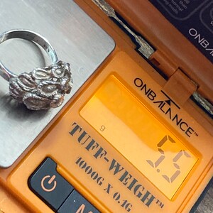 May include: A silver ring with a textured design is weighed on a digital scale. The scale reads 5.5 grams. The scale is orange and black and says "TUFF-WEIGH&trade; 1000G X 0.1G" on it.
