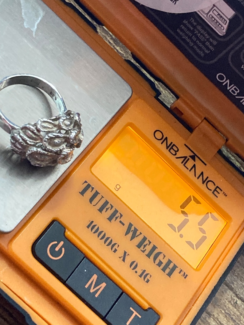 May include: A silver ring with a textured design is weighed on a digital scale. The scale reads 5.5 grams. The scale is orange and black and says "TUFF-WEIGH&trade; 1000G X 0.1G" on it.