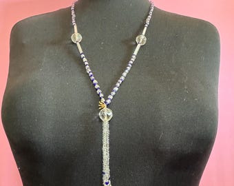 May include: A Y-shaped beaded necklace. It features clear and blue beads, clear glass orbs, and silver-coloured metal tubes. A clear, beaded tassel hangs from the centre, with gold-coloured accents.