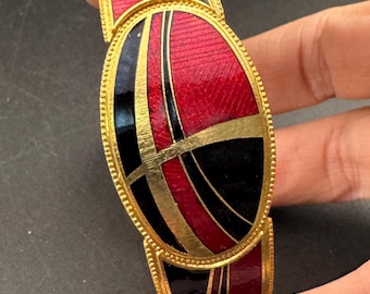 May include: Gold-toned metal bracelet with red, black, and gold enamel design. The bracelet features an oval centerpiece and rectangular links, with a textured pattern on the red enamel.