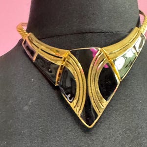 May include: A gold-toned statement necklace with black enamel accents. The necklace features a geometric design with a V-shaped pendant and a textured chain. The pendant has a diamond shape in the center.