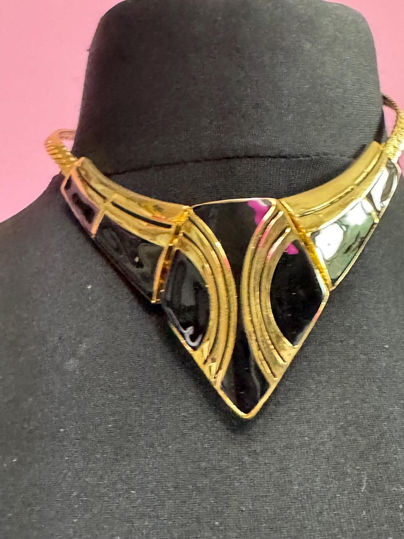 May include: A gold-toned statement necklace with black enamel accents. The necklace features a geometric design with a V-shaped pendant and a textured chain. The pendant has a diamond shape in the center.