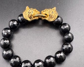 May include: A bracelet featuring a series of round, faceted black beads arranged in a circular pattern. The two ends of the bead strand are connected by a gold-toned metal clasp designed as two facing tiger heads joined by a small ring.