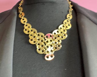 May include: A gold-colored statement necklace with a unique design. The necklace features interconnected circular and oval shapes, creating a layered effect. The jewelry is displayed on a black mannequin, with a pink background.