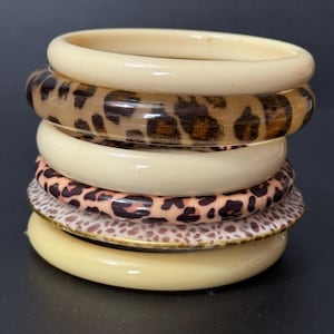 Peut inclure: Un ensemble de six bracelets en plastique, empilés les uns sur les autres. Les bracelets sont de couleur crème et présentent un motif léopard.