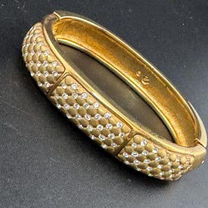 May include: Gold-toned hinged bracelet with a textured surface and small, clear rhinestones. The bracelet has a rectangular shape and is set against a dark background. The bracelet is a piece of jewelry.