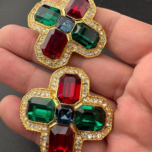 May include: A pair of gold tone earrings with a cross design. The earrings feature red, green, and blue gemstones.