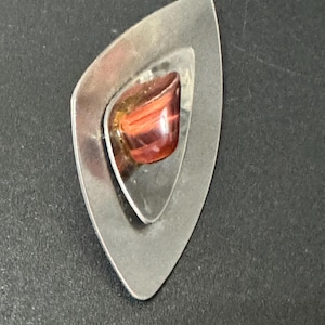 May include: A silver pendant with a geometric design featuring a red and orange stone.
