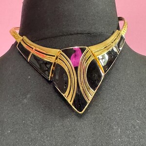 May include: A gold-toned necklace with a geometric design. The necklace features black enamel accents and a central diamond-shaped pendant with a pink detail. The necklace is displayed on a black mannequin head against a pink background.