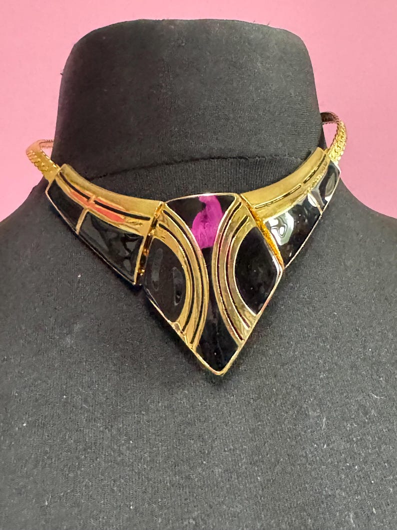 May include: A gold-toned necklace with a geometric design. The necklace features black enamel accents and a central diamond-shaped pendant with a pink detail. The necklace is displayed on a black mannequin head against a pink background.