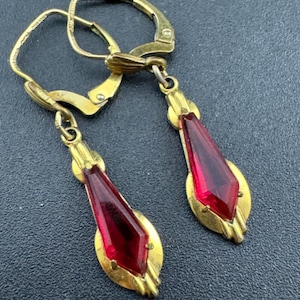 以下が含まれることがあります： レバーバック留め金が付いた、ヴィンテージのゴールドトーンのイヤリングのペア。各イヤリングは、ルビーレッドの細長いダイヤモンド型のガラスストーンを、ゴールドトーンの金属フレームにセットしています。イヤリングはクラシックなアールデコデザインです。