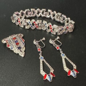 May include: A vintage jewellery set featuring a silver tone bracelet, brooch, and earrings. The set is decorated with clear, red, and blue rhinestones in a geometric pattern.