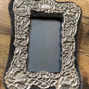 Puede incluir: Un marco de fotos de plata con un dise&ntilde;o ornamentado y un respaldo de terciopelo negro. El marco tiene una abertura rectangular para una foto y est&aacute; marcado con "KEID" y "925" en la parte inferior.