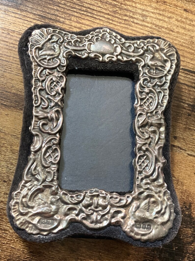 Puede incluir: Un marco de fotos de plata con un dise&ntilde;o ornamentado y un respaldo de terciopelo negro. El marco tiene una abertura rectangular para una foto y est&aacute; marcado con "KEID" y "925" en la parte inferior.