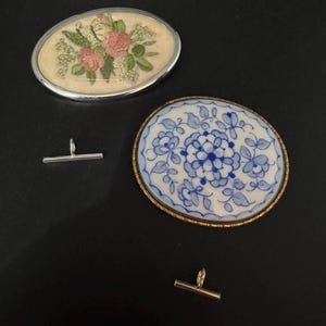May include: Two oval brooches and two tie bars on a black surface. One brooch has a silver frame and features floral embroidery with pink roses. The other brooch has a gold frame and a blue and white floral design. The tie bars are silver and gold.
