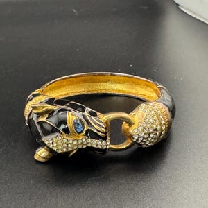 以下が含まれることがあります： 黒いパンサーの頭部をモチーフにしたヒンジ付きバングルブレスレット。ゴールドトーンの金属アクセント、鼻先と反対側の端に施されたクリアラインストーンのパヴェ装飾、目には小さな青い石がはめ込まれており、内側は光沢のあるゴールドトーン仕上げになっています。