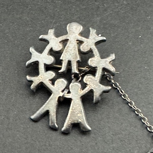 May include: A silver brooch depicting a circle of stylized figures holding hands. The figures are arranged around a central figure, with a chain attached. The brooch has a simple, elegant design.