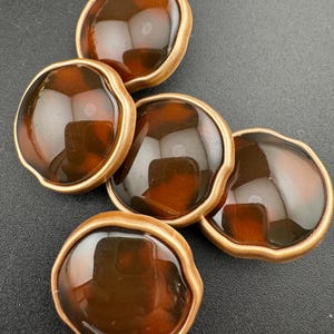 May include: A collection of six brown and gold buttons. Each button has a rounded, irregular shape with a glossy, tortoiseshell-like center and a gold-colored rim. The buttons are arranged on a dark surface.