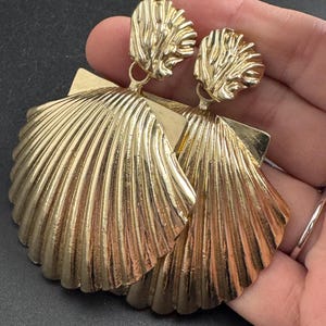 May include: A pair of gold-coloured seashell earrings. The earrings feature a textured, ridged design, with a square backing and a smaller, textured top piece. The earrings are made of a shiny, metallic material.