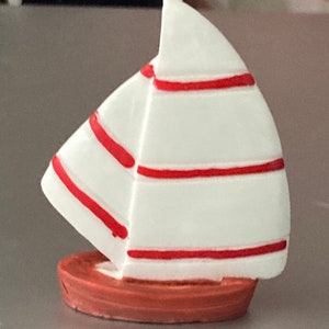 以下が含まれることがあります： 赤いストライプのついた小さな白い帆船。帆船はプラスチック製で、茶色の土台があります。