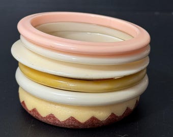 以下が含まれることがあります： ピンク、白、黄色のプラスチック製のバングルが5つ重ねられています。一番下のバングルには、茶色のジグザグ模様があります。