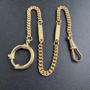 May include: A gold-coloured pocket watch chain with a circular clasp and a spring-loaded hook. The chain features a curb link design with decorative rectangular links. The chain is displayed on a dark surface.
