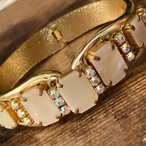 May include: A gold bangle bracelet with a row of five rectangular white mother-of-pearl stones set in gold. Each stone is accented with clear rhinestones.