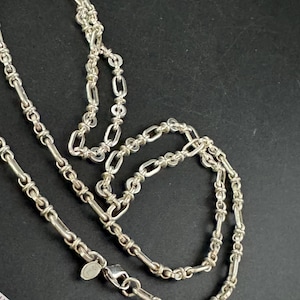 May include: A silver necklace featuring a mix of chain link designs. The necklace, made from a shiny metal, has a lobster clasp. It is presented against a dark backdrop.