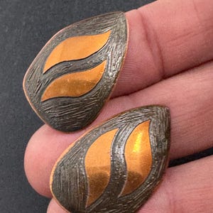 May include: A pair of copper-coloured earrings with a textured, dark grey background and two orange-gold leaf-shaped inlays. The earrings are teardrop-shaped and have a unique, handcrafted appearance.