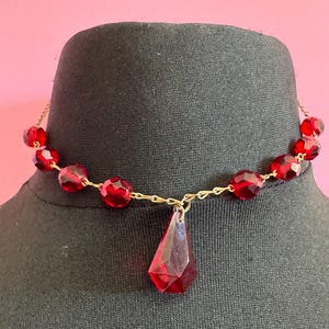May include: A red beaded necklace with a gold chain. The necklace features faceted red beads and a large, teardrop-shaped pendant. The necklace is displayed on a black mannequin head against a pink background.