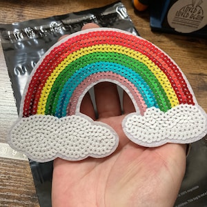 May include: A rainbow-coloured sequin patch with white clouds. The rainbow is made of red, orange, yellow, green, blue, and pink sequins. The clouds are made of white sequins.