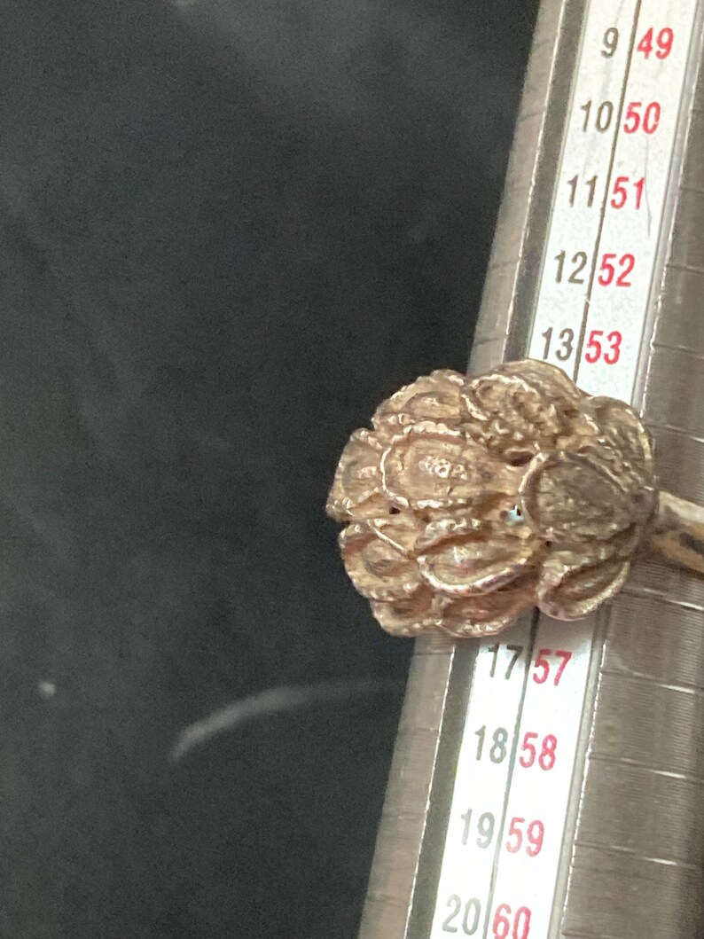 May include: A silver ring with a detailed floral design. The ring is being measured with a ring sizer, showing a size of 17.5.