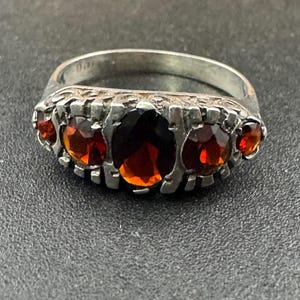 以下が含まれることがあります： 5つのオレンジがかった赤色の宝石が付いたシルバーリング。中央の石は楕円形で、両側に2つの小さな丸い石が配置されています。リングバンドはテクスチャード加工のデザインです。