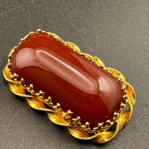 May include: A rectangular, reddish-brown gemstone brooch with a gold-colored filigree border. The gemstone has a polished, glossy surface. The brooch has a decorative, vintage design.