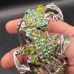 May include: Silver frog-shaped bracelet adorned with iridescent green and yellow rhinestones. The bracelet showcases a detailed frog design with textured legs and body. The rhinestones add sparkle and colour to the jewellery.