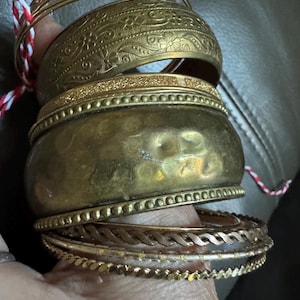 May include: A selection of gold-coloured bangles. The bangles feature diverse designs, such as floral patterns, hammered textures, and beaded edges. Some are slender, while others are broader. A red and white string is visible.