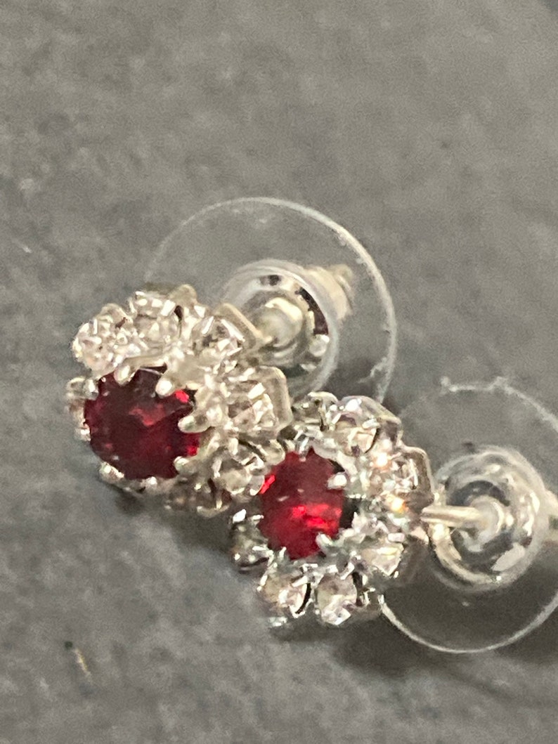 Puede incluir: Un par de pendientes de plata con una piedra roja rodeada de piedras blancas m&aacute;s peque&ntilde;as.
