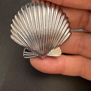 May include: A silver-colored seashell-shaped brooch. The brooch has a detailed, ridged design, with a fan-like structure. The metal has a shiny, reflective surface. The brooch is a decorative accessory.