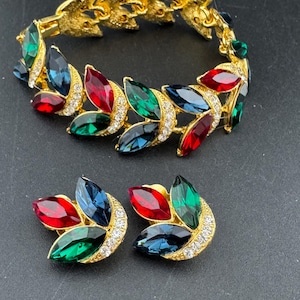 May include: A gold-coloured jewellery set comprising a bracelet and matching earrings. The bracelet and earrings are decorated with red, green, and blue jewel-toned stones, with small clear crystals. The jewellery has a leaf-like design.