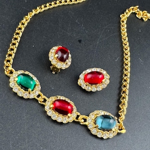 May include: A gold-coloured necklace and earring set. The necklace has a chain design with oval-shaped gemstones in red, green, and blue, each surrounded by small, clear crystals. The earrings mirror the necklace design, with a red gemstone and crystal halo.