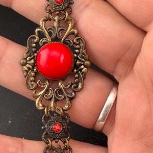May include: A gold-tone bracelet with a large red cabochon stone surrounded by intricate filigree details. The bracelet is adorned with smaller red rhinestones.