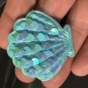 May include: A light blue seashell-shaped patch with iridescent sequins. The patch is made of fabric and has a raised, textured design.