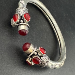 May include: A silver bracelet with intricate patterns and red gemstone embellishments. The bracelet has a curved shape with decorative ends, each featuring several red stones. The design suggests a vintage or ethnic style, with a focus on detail.